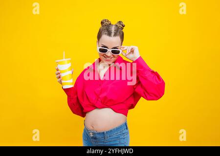 Eine süße schwangere Frau in einem rosa Hemd trinkt Wasser aus einem Plastikbecher. Eine schwangere Frau auf gelbem Hintergrund mit einem Glas in der Hand. Schädliches Wasser Stockfoto