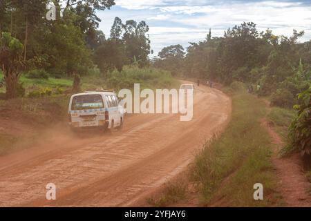 Ein Taxibus, auch bekannt als „Matatu“, fährt auf einer rötlichen Feldstraße in einer ländlichen Gegend Ugandas, umgeben von üppigem Grün. Stockfoto