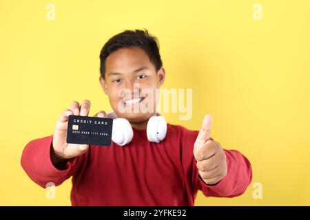 Übergewichtiger asiatischer Junge, der Black Card hält, während er Musik hört, isoliert auf gelbem Hintergrund Stockfoto