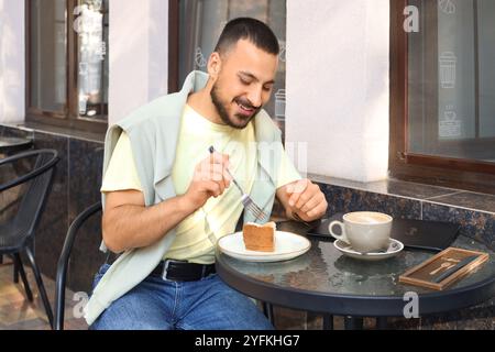 Junger Mann mit Menü, der Kuchen am Tisch im Straßencafé isst Stockfoto