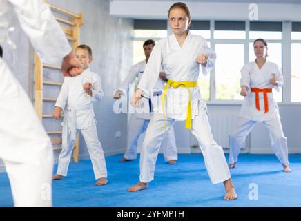 Teenager-Mädchen in weißem Kimono gekleidet mit Gürtel-Übungstechniken mit Trainer, im Hintergrund macht ihre Familie Kata Stockfoto