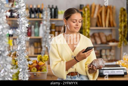 Die junge Frau im Geschäft wählt gesaugten Schweineschinken, scannt das Produktetikett mit dem Telefon Stockfoto