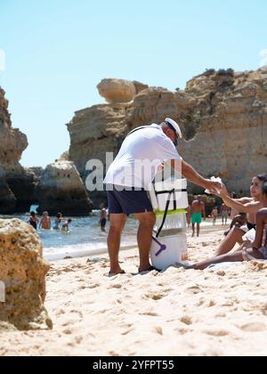 Ein freundlicher Verkäufer verkauft eine traditionelle Bola de Berlim an einen Strandgänger im Praia de São Rafael, eingerahmt von den Klippen der Algarve Stockfoto