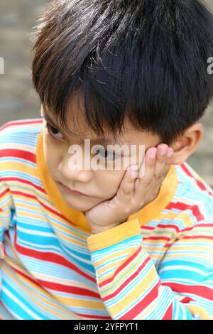 Ein nachdenklicher kleiner Junge in einem bunten gestreiften Hemd zeigt einen nachdenklichen Ausdruck, der sein Kinn auf seiner Hand hält. Diese Nahaufnahme fängt die unschuldige Kindheit ein Stockfoto