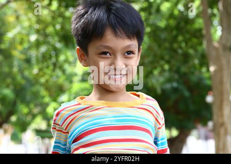 Ein fröhlicher kleiner Junge lächelt in einem bunten gestreiften Hemd. Umgeben von lebendigem Grün verkörpert er Glück und unbeschwertes Kinderhoo Stockfoto