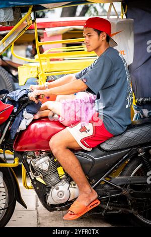 Ein junger philippinischer Mann sitzt auf seinem Trike im Verkehr, und seine kleine Tochter saß auf seinem Schoß in Manila City, den Philippinen. Stockfoto