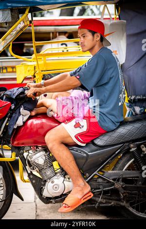 Ein junger philippinischer Mann sitzt auf seinem Trike im Verkehr, und seine kleine Tochter saß auf seinem Schoß in Manila City, den Philippinen. Stockfoto