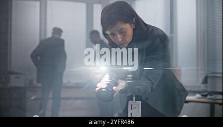 Büroraub oder Mordfall: Eine forensische Wissenschaftlerin fotografiert Beweisaufnahmen mit Kamera. Afroamerikanischer Polizist und Detective im Hintergrund. Das Ermittlungsteam arbeitet am Tatort. Stockfoto