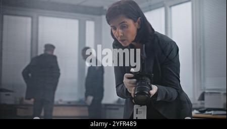 Büroraub oder Mordfall: Eine forensische Wissenschaftlerin fotografiert Beweisaufnahmen mit Kamera. Afroamerikanischer Polizist und Detective im Hintergrund. Das Ermittlungsteam arbeitet am Tatort. Stockfoto
