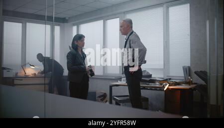 Büroraub oder Mordfall: Ermittlungsteam am Tatort. Die weibliche Forensikologin fotografiert Beweise und spricht dann mit einem männlichen Detektiv. Der afroamerikanische Polizist sucht nach Fingerabdrücken. Stockfoto