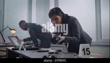Ermittlungsteam am Tatort: Kriminalistin fotografiert Hinweise, afroamerikanischer Polizist durchsucht Fingerabdrücke. Nummerierte Markierung neben Beweis. Büroraub oder Mordfall. Stockfoto