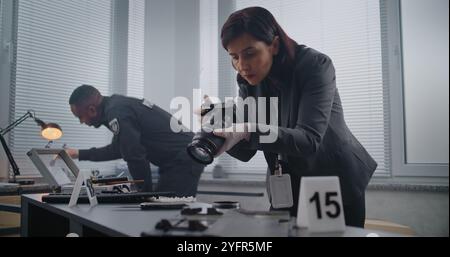 Ermittlungsteam am Tatort: Kriminalistin fotografiert Hinweise, afroamerikanischer Polizist durchsucht Fingerabdrücke. Nummerierte Markierung neben Beweis. Büroraub oder Mordfall. Stockfoto