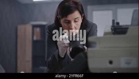 Büroraub oder Mordfall: Eine Kriminalistin fotografiert Beweise am Tatort mit Kamera. Afroamerikanischer Polizist und Detective im Hintergrund. Das Untersuchungsteam arbeitet. Stockfoto