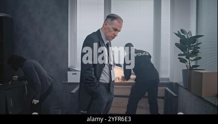 Büroraub oder Mordfall: Kriminalermittlungsteam arbeitet. Ein männlicher Detective untersucht Beweise am Tatort, Kriminalistin und afroamerikanische Polizistin suchen nach Fingerabdrücken. Stockfoto