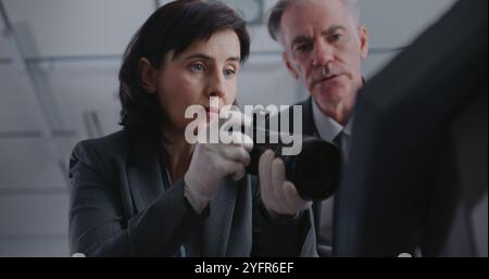 Büroraub oder Mordfall: Nahaufnahme einer Kriminalistin, die mit einer professionellen Kamera Beweisaufnahmen am Tatort macht. Ein männlicher Detective kommt, spricht mit einem Kollegen. Das Untersuchungsteam arbeitet. Stockfoto
