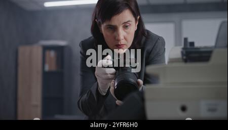 Büroraub oder Mordfall: Eine Kriminalistin fotografiert Beweise am Tatort mit Kamera. Afroamerikanischer Polizist und Detective im Hintergrund. Das Untersuchungsteam arbeitet. Stockfoto