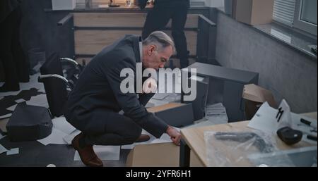 Büroraub oder Mordfall: Kriminalermittlungsteam arbeitet. Ein männlicher Detective untersucht Beweise am Tatort, Kriminalistin und afroamerikanische Polizistin suchen nach Fingerabdrücken. Stockfoto