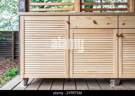 Holzschränke der Außenküche mit Lamellen, Schubladen, Holzgriffen und Sperrholzplatte stehen auf einer Holzterrasse Stockfoto