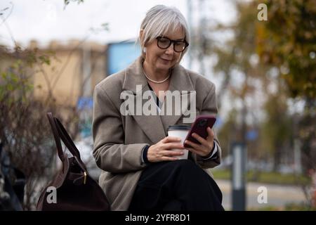 Eine ältere Frau genießt einen Kaffee und benutzt ihr Telefon, während sie an einem sonnigen Herbsttag auf einer Bank im Park sitzt Stockfoto