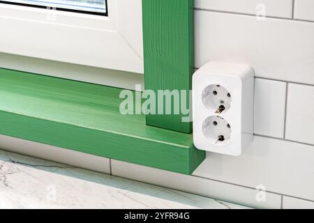 Fragment eines Kunststofffensters mit grüner hölzerner Fensterbank und Holzrahmen mit Steckdose an der gefliesten Wand aus der Nähe Stockfoto