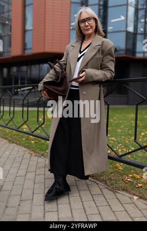 Eine ältere Geschäftsfrau mit stilvollem Mantel steht im Herbst selbstbewusst vor ihrem Bürogebäude Stockfoto