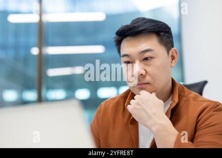 Asiatischer Mann Geschäftsmann im Büro, der nachdenklich am Laptop arbeitet, sich auf Aufgaben und Ideen in modernem Ambiente konzentriert und Geschäfts- und Technologiekonzepte präsentiert Stockfoto