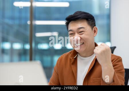 Asiatischer Geschäftsmann, der im modernen Büro sitzt und fröhlich lächelt, mit Faustpumpe Erfolg macht, während er auf dem Laptop arbeitet, was Leistung, Positivität und professionelle Zufriedenheit vermittelt. Stockfoto