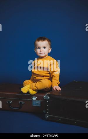 Ein kleiner Junge von 1 Jahr in einem gelben Strickanzug auf blauem Hintergrund. Alltagsgegenstände für Kinder. Stockfoto