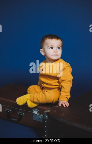 Ein kleiner Junge von 1 Jahr in einem gelben Strickanzug auf blauem Hintergrund. Alltagsgegenstände für Kinder. Stockfoto