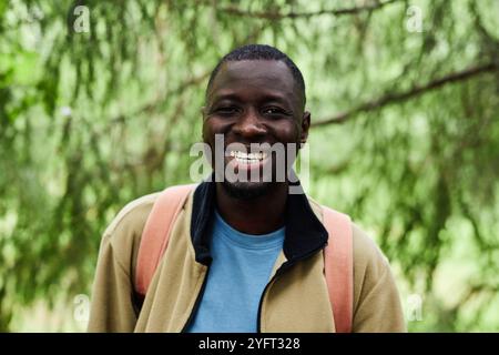 Vorderansicht Porträt eines afroamerikanischen Mannes, der fröhlich in die Kamera lächelt, während er mit dem Rucksack im grünen Wald wandert Stockfoto