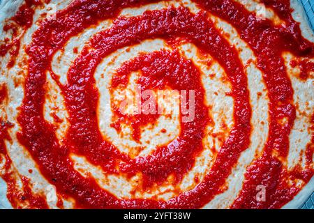 Ein Kreis Pizzateig ist mit Tomatensauce in Form einer Spirale bedeckt. Der Prozess der Pizza. Hintergrund mit der Textur von Teig und roter Soße. Nahaufnahme, Draufsicht. Hochwertige Fotos Stockfoto