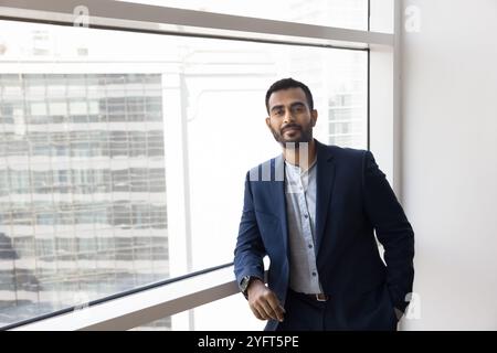 Positiver, selbstbewusster junger indischer Manager, der am Fenster des Büros steht Stockfoto