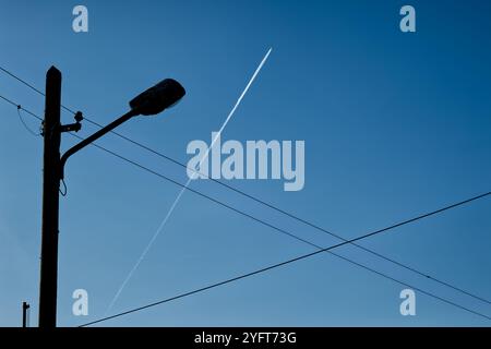 Strommasten mit Lampenpfosten und überquerenden Stromleitungen gegen einen klaren blauen Himmel und ein einzelnes Flugzeug mit einer geraden weißen Kondensationsspur Stockfoto