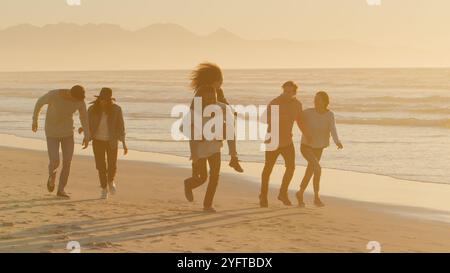 Mann Gibt Frau Huckepack Als Gruppe Lässig Gekleideter Freunde, Die Zusammen Am Winter Beach Laufen Stockfoto