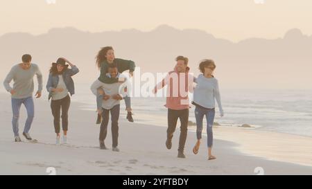 Mann Gibt Frau Huckepack Als Gruppe Lässig Gekleideter Freunde, Die Zusammen Am Winter Beach Laufen Stockfoto