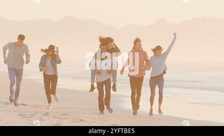 Mann Gibt Frau Huckepack Als Gruppe Lässig Gekleideter Freunde, Die Zusammen Am Winter Beach Laufen Stockfoto