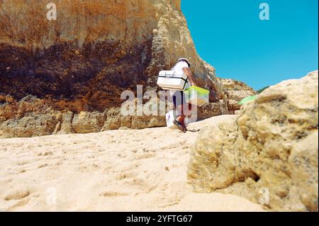 Ein engagierter, ausgereifter Anbieter klettert auf die Sandhänge des Praia de São Rafael, während er traditionelle Bolas de berlim in Styropor-Kühlern mitbringt. Stockfoto