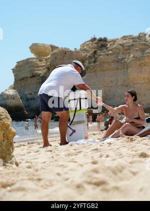 Ein freundlicher Verkäufer verkauft eine traditionelle Bola de Berlim an einen Strandgänger im Praia de São Rafael, eingerahmt von den Klippen der Algarve Stockfoto