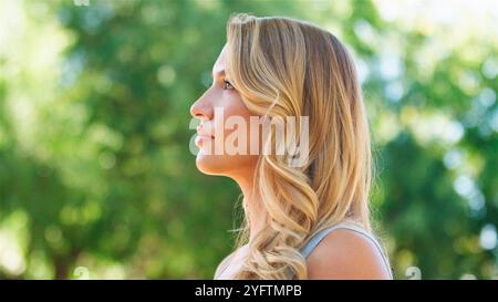 Eine Frau steht im Profil und blickt nachdenklich in die Ferne, umgeben von lebendigem Grün. Das Sonnenlicht filtert durch die Blätter, Castin Stockfoto