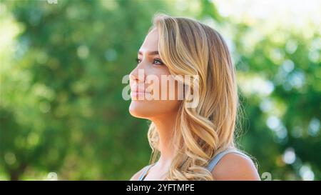 Eine Frau steht draußen und lächelt sanft, während sie in den klaren blauen Himmel blickt. Umgeben von üppig grünen Bäumen, betont das Sonnenlicht ihre Gesichtszüge. Stockfoto