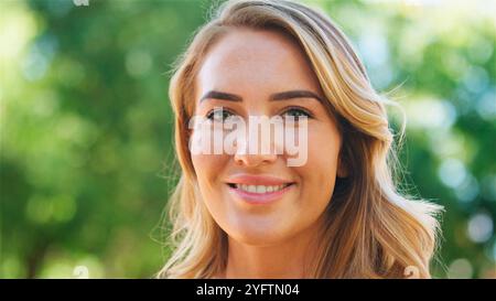 Eine junge Frau steht draußen mit einem warmen, einladenden Lächeln, umgeben von üppigem Grün. Das Sonnenlicht zieht durch die Blätter und hebt sie fröhlich hervor Stockfoto