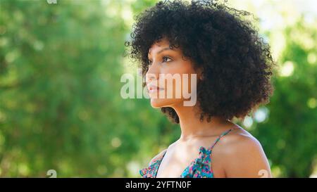 Eine junge Frau steht anmutig draußen und zeigt ihr lockiges Haar und ihre leuchtende Kleidung. Sonnenlicht filtert durch das üppige Grün, das sie umgibt, cr Stockfoto