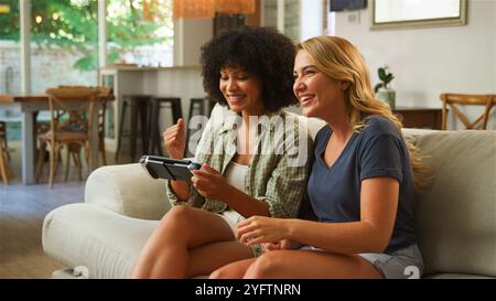 Zwei Freunde sitzen bequem auf einem Sofa in einem hellen Wohnzimmer und teilen sich Lachen und Aufregung, während sie gemeinsam ein Videospiel auf einer Handhel spielen Stockfoto