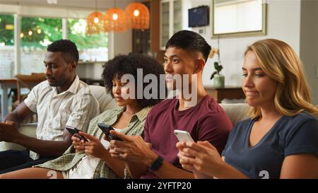 Eine bunte Gruppe von vier Freunden sitzt zusammen auf einer Couch in einem hellen, einladenden Wohnzimmer. Jede Person konzentriert sich auf ihr Smartphone, scheint es Stockfoto