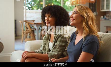 Zwei junge Frauen sitzen bequem auf einer Couch in einem hellen Wohnzimmer und unterhalten sich lebhaft. Die entspannte Atmosphäre bietet warme Beleuchtung und Stockfoto