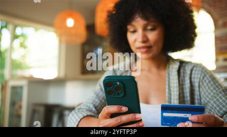 Eine junge Frau sitzt in einem gemütlichen Innenbereich und nutzt ihr Smartphone, während sie eine Kreditkarte hält. Der Raum ist warm beleuchtet mit Hängeleuchte Stockfoto