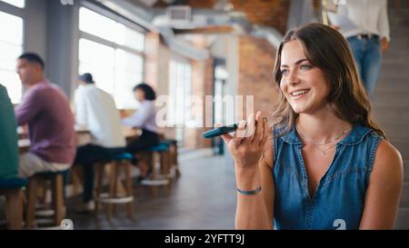 Geschäftsfrau, Die Im Open Plan Office Mit Kollegen Im Hintergrund Am Mobiltelefon Mit Dem Mikrofon Spricht Stockfoto