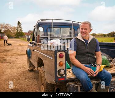 Reifer Männlicher Landarbeiter Sitzt Auf Der Heckklappe Eines Geländefahrzeugs Und Hält Heißgetränk Im Cup Stockfoto