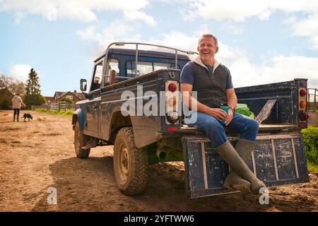 Reifer Männlicher Landarbeiter Sitzt Auf Der Heckklappe Eines Geländefahrzeugs Und Hält Heißgetränk Im Cup Stockfoto