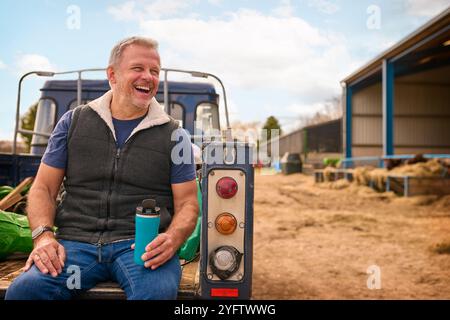 Reifer Männlicher Landarbeiter Sitzt Auf Der Heckklappe Eines Geländefahrzeugs Und Hält Heißgetränk Im Cup Stockfoto